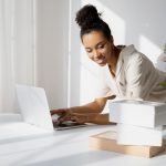African American woman working on a laptop with boxes and flowers in a bright room.