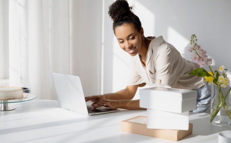 African American woman working on a laptop with boxes and flowers in a bright room.